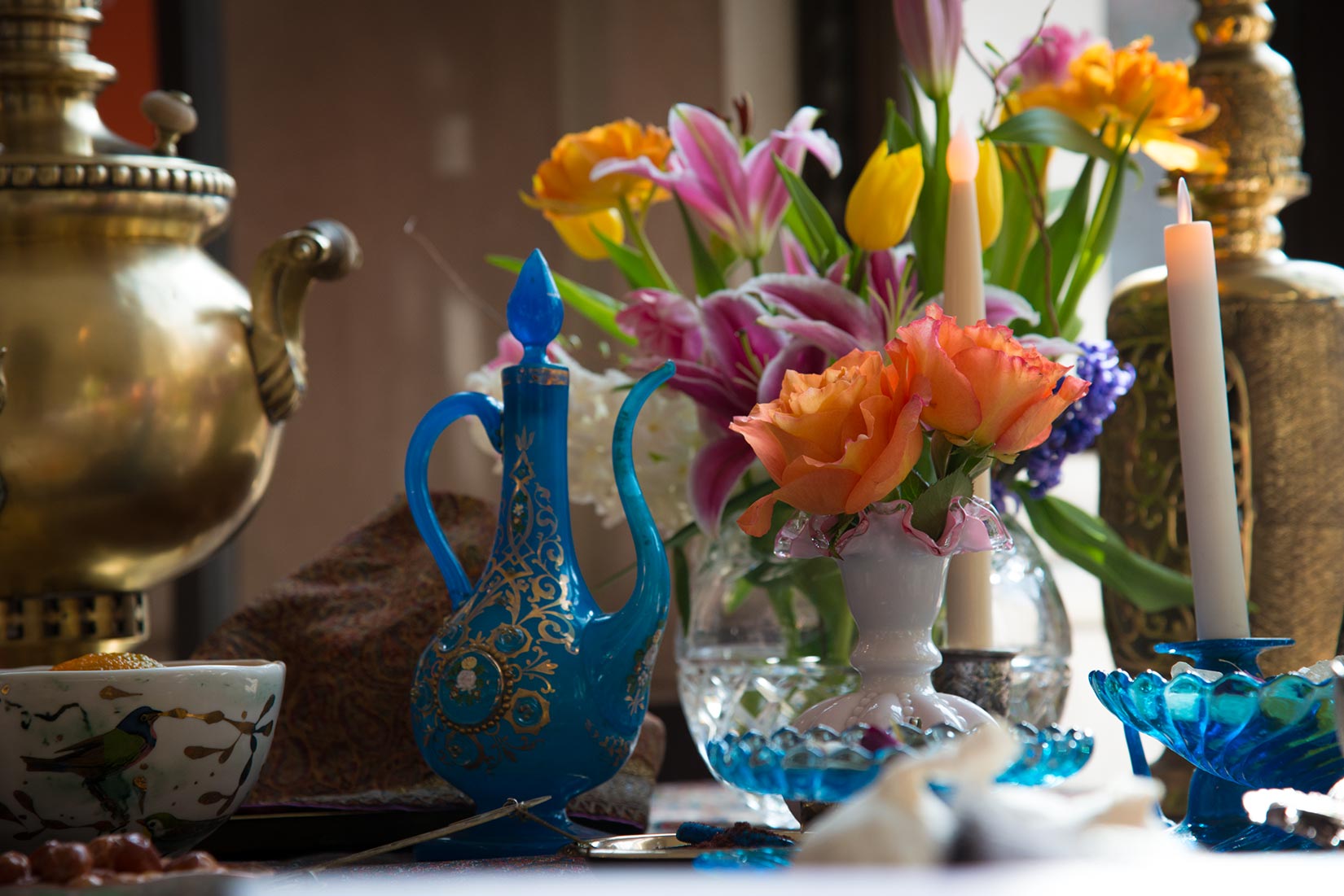 A table, viewed close up, that's been set with a vase of flowers, a blue-and-gold spouted jar, and candles.